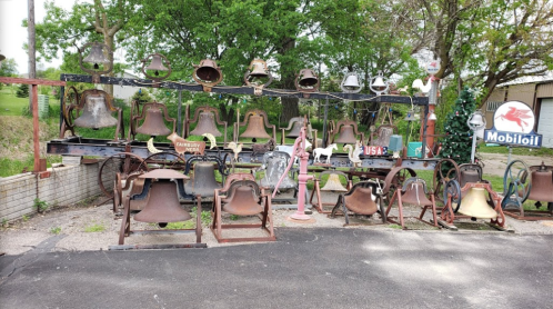 A collection of various antique bells displayed on a rack, with trees and a Mobil oil sign in the background.