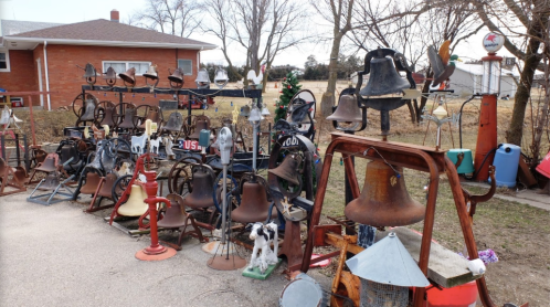 A collection of various bells displayed outdoors, with a house and trees in the background.