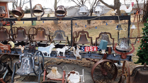 A collection of vintage bells and metal horse-drawn equipment displayed outdoors, with a festive touch in the background.