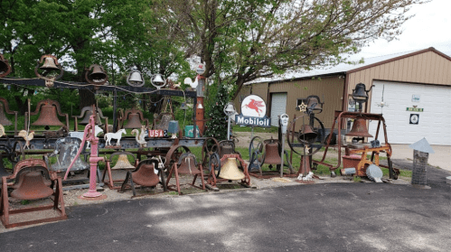 A collection of various bells and vintage items displayed outdoors, with trees and a building in the background.