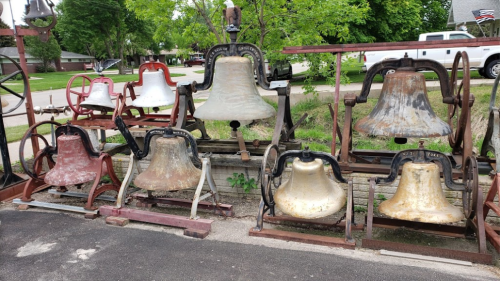 A collection of various large bells displayed outdoors, with different colors and sizes, set against a green backdrop.