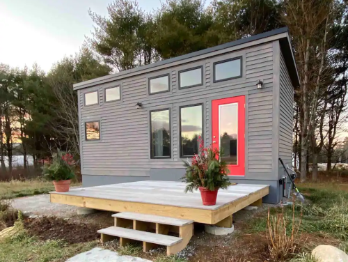 A modern tiny house with a red door, large windows, and a wooden deck, surrounded by trees and a grassy area.