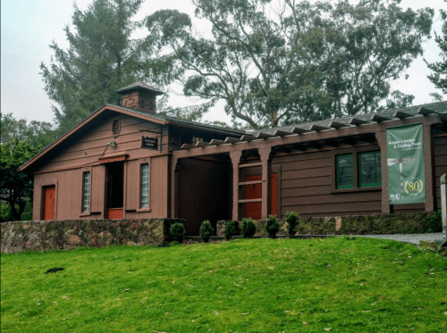 A rustic wooden building with a sloped roof, surrounded by greenery and featuring a banner on the side.