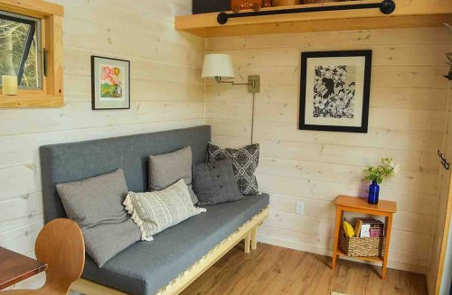 Cozy interior of a tiny home featuring a gray sofa, wooden accents, artwork, and a small side table with decor.