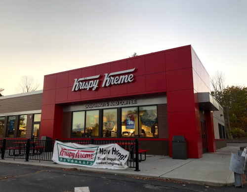 Exterior of a Krispy Kreme store with a red facade and a hiring banner in front.