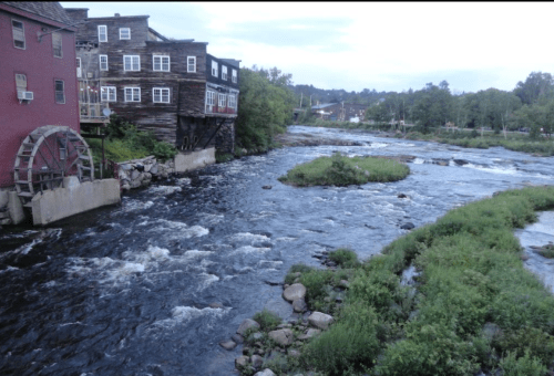 A river flows past a wooden building with a waterwheel, surrounded by greenery and rocky banks under a cloudy sky.