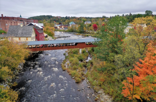 A scenic view of a covered bridge over a river, surrounded by colorful autumn foliage and quaint buildings.