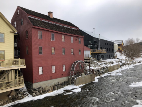 A red mill building with a water wheel beside a river, surrounded by snow and other buildings in a winter landscape.