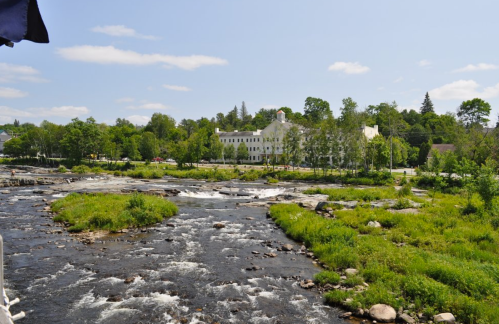 A scenic view of a river with rocky banks, lush greenery, and a white building in the background under a clear blue sky.