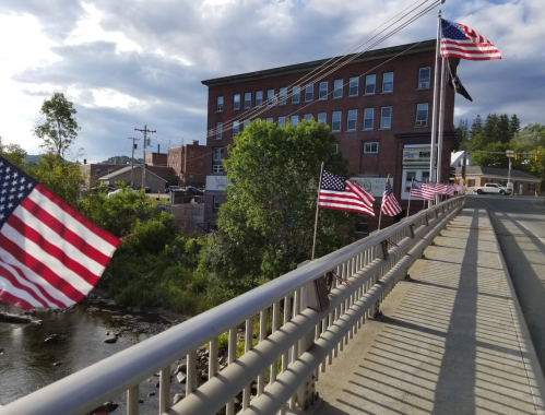 A bridge lined with American flags, overlooking a river and a historic building under a cloudy sky.
