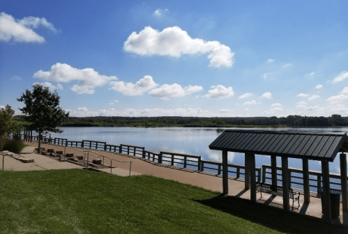 A serene lakeside view with a wooden walkway, benches, and a gazebo under a clear blue sky with fluffy clouds.