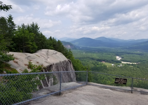 A scenic view from a rocky overlook, surrounded by trees and mountains under a cloudy sky. A warning sign is visible.
