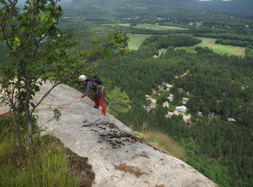 A climber in a helmet ascends a rocky cliff, overlooking a lush green valley and small houses below.