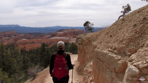 A hiker with a red backpack walks along a rocky trail, surrounded by colorful cliffs and trees in a scenic landscape.