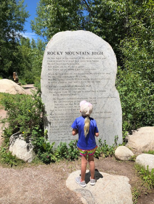 A child in a blue shirt and colorful shorts stands in front of a large stone with the text of "Rocky Mountain High."