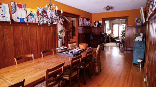 A warm-toned dining room with a wooden table, chairs, and colorful artwork on the walls, leading to a cozy living area.