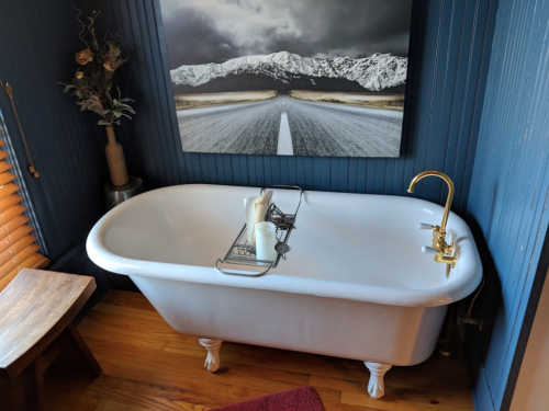 A vintage white bathtub with a gold faucet, a candle, and a scenic mountain road painting on the wall.