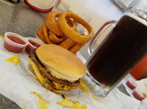 A burger with melted cheese, onion rings, and a large mug of dark soda, served with ketchup on a diner-style table.