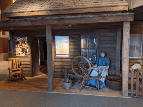 A wooden cabin display with a spinning wheel, featuring a seated woman and a boy beside her.