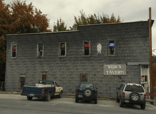 A weathered gray building with neon signs for a tavern, featuring parked trucks in front and autumn foliage in the background.