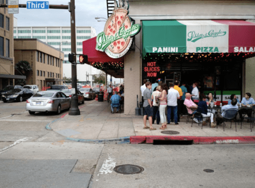 A busy street corner with a pizza restaurant, outdoor seating, and people dining on the sidewalk.