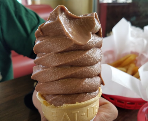 A hand holding a soft-serve chocolate ice cream cone with a textured swirl, set against a background of fries.