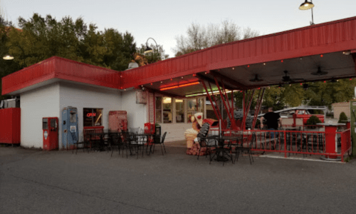 A red and white fast-food restaurant with outdoor seating, illuminated by string lights, and a vintage soda machine.