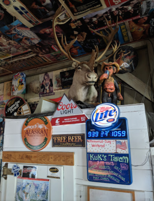 A rustic bar scene featuring a mounted moose head, neon signs, and various beer advertisements on the walls.