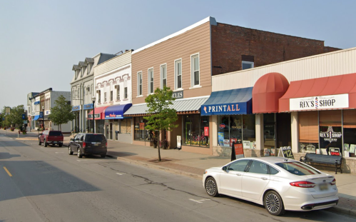 A street view of shops with awnings, parked cars, and trees lining the sidewalk on a sunny day.