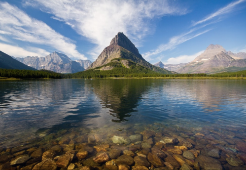 A serene lake reflects a towering mountain under a blue sky with wispy clouds, surrounded by lush greenery.