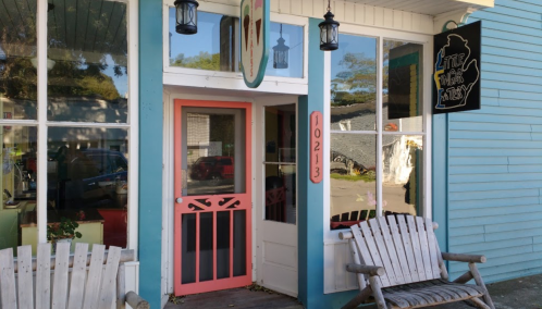 A charming storefront with a pink door, wooden chairs, and colorful signage, set in a quaint neighborhood.