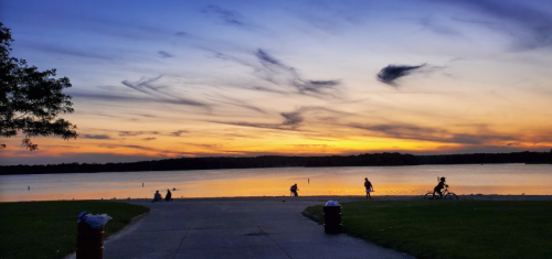 A serene sunset over a lake, with silhouettes of people walking and biking along the shore.