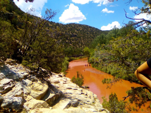 A scenic view of a reddish river winding through green trees and rocky terrain under a blue sky with fluffy clouds.