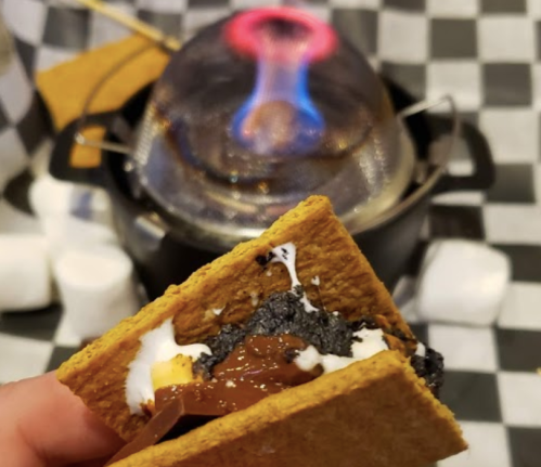 A hand holds a s'more with melted chocolate and marshmallow, with a flaming dessert maker in the background.