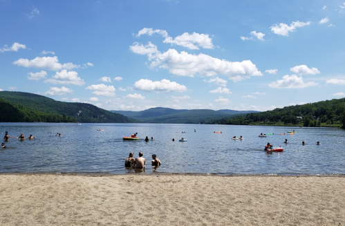 A sandy beach by a lake with people swimming and enjoying the sun under a blue sky with fluffy clouds.