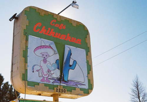 A colorful sign for "Café Chihuahua" featuring a cartoon dog in a sombrero next to a cactus, with some damage visible.