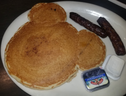 A large pancake shaped like a bear, accompanied by two sausage links and a small container of syrup.