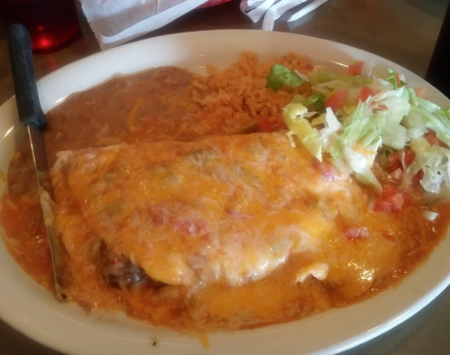 A plate of enchiladas topped with melted cheese, served with rice, beans, and a side of lettuce and tomatoes.
