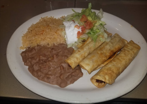 A plate with three crispy taquitos, refried beans, rice, and a side salad topped with tomatoes and sour cream.