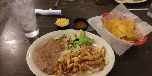 A plate of tacos with chicken, refried beans, salad, lime, cheese, salsa, and a side of tortilla chips.