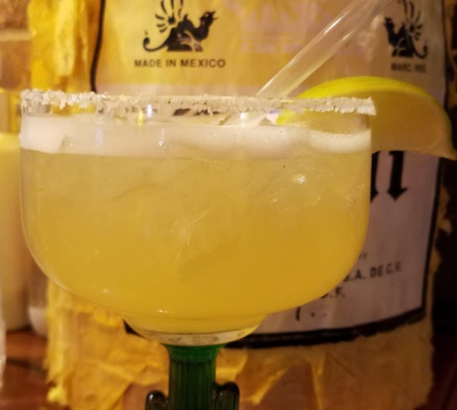 A close-up of a margarita in a glass with a salted rim, garnished with a lemon slice, set against a rustic background.