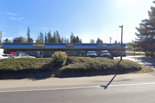 A row of storefronts with various signs, surrounded by greenery and parked cars, under a clear blue sky.