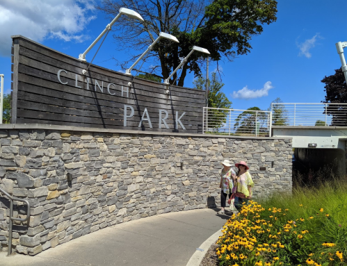 Two people walk near a stone wall with a sign that reads "Clinch Park" under a blue sky with scattered clouds.