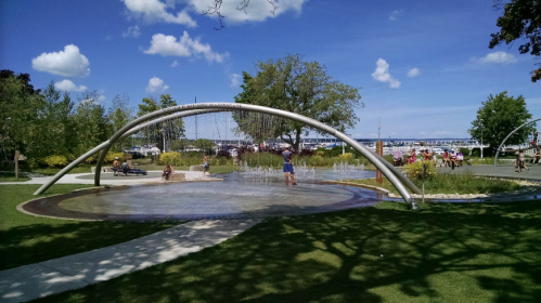 A sunny park scene with a water feature, trees, and people enjoying the outdoors near a waterfront.