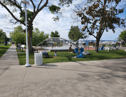 A park scene with children playing near a water fountain, trees, and people enjoying the outdoors on a sunny day.
