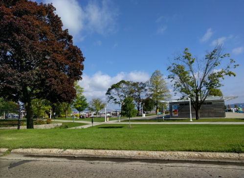 A park scene with green grass, trees, and a building under a blue sky with clouds. People are visible in the background.