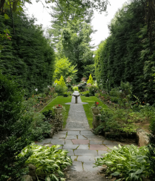 A serene garden path lined with greenery, leading to a central stone feature amidst blooming flowers.