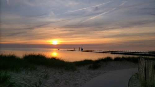 A serene beach at sunset, with soft waves, a pier in the distance, and wispy clouds in a colorful sky.