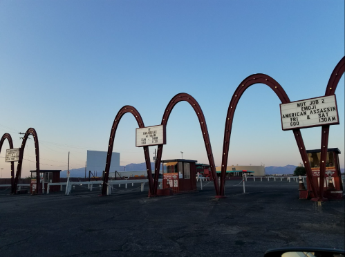 Drive-in movie theater entrance with arched structures and signage displaying movie titles and showtimes at dusk.