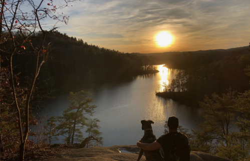 A person and a dog sit on a rock, watching a sunset over a calm lake surrounded by trees.